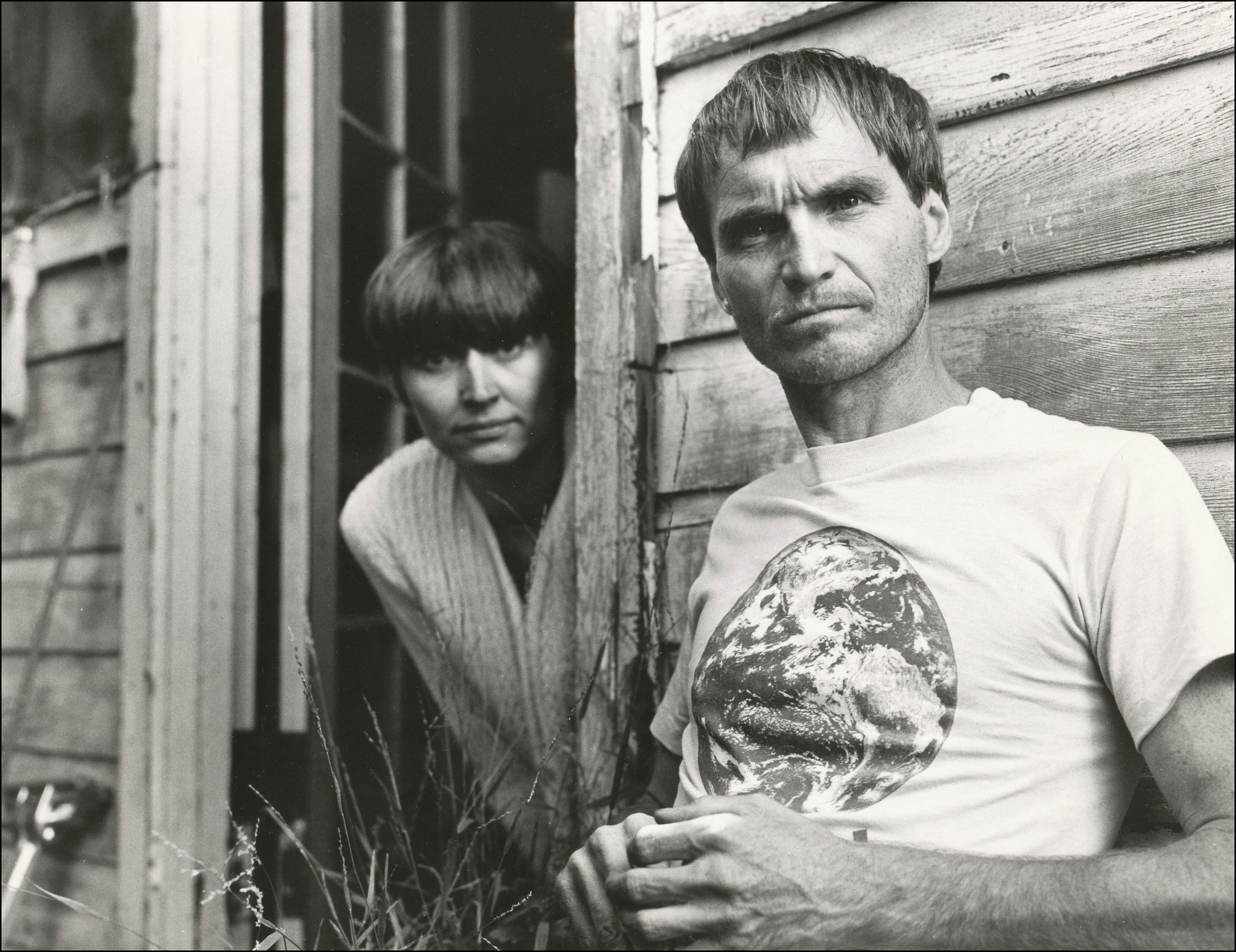 Man and woman looking at camera. Man in focus in front of wood house. Man wearing t-shirt with picture of the earth. Woman blurred in background looking out from door