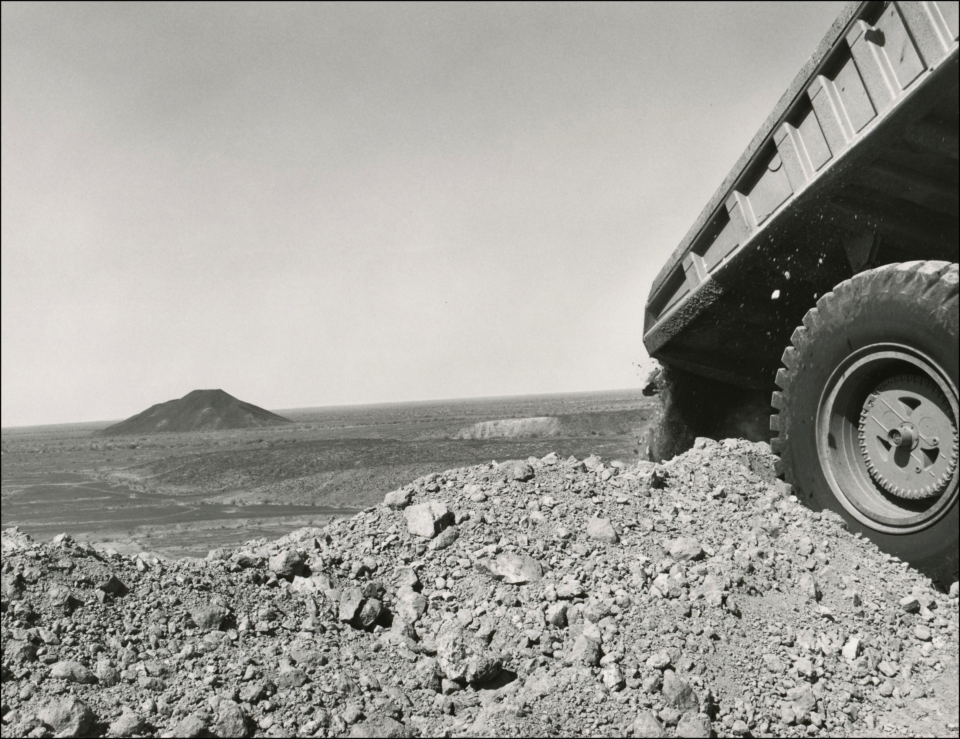 View of the back corner of a dump truck dumping dirt and rock in a pile at a gold mine. Flat open space with one cone shaped hill in the background distance
