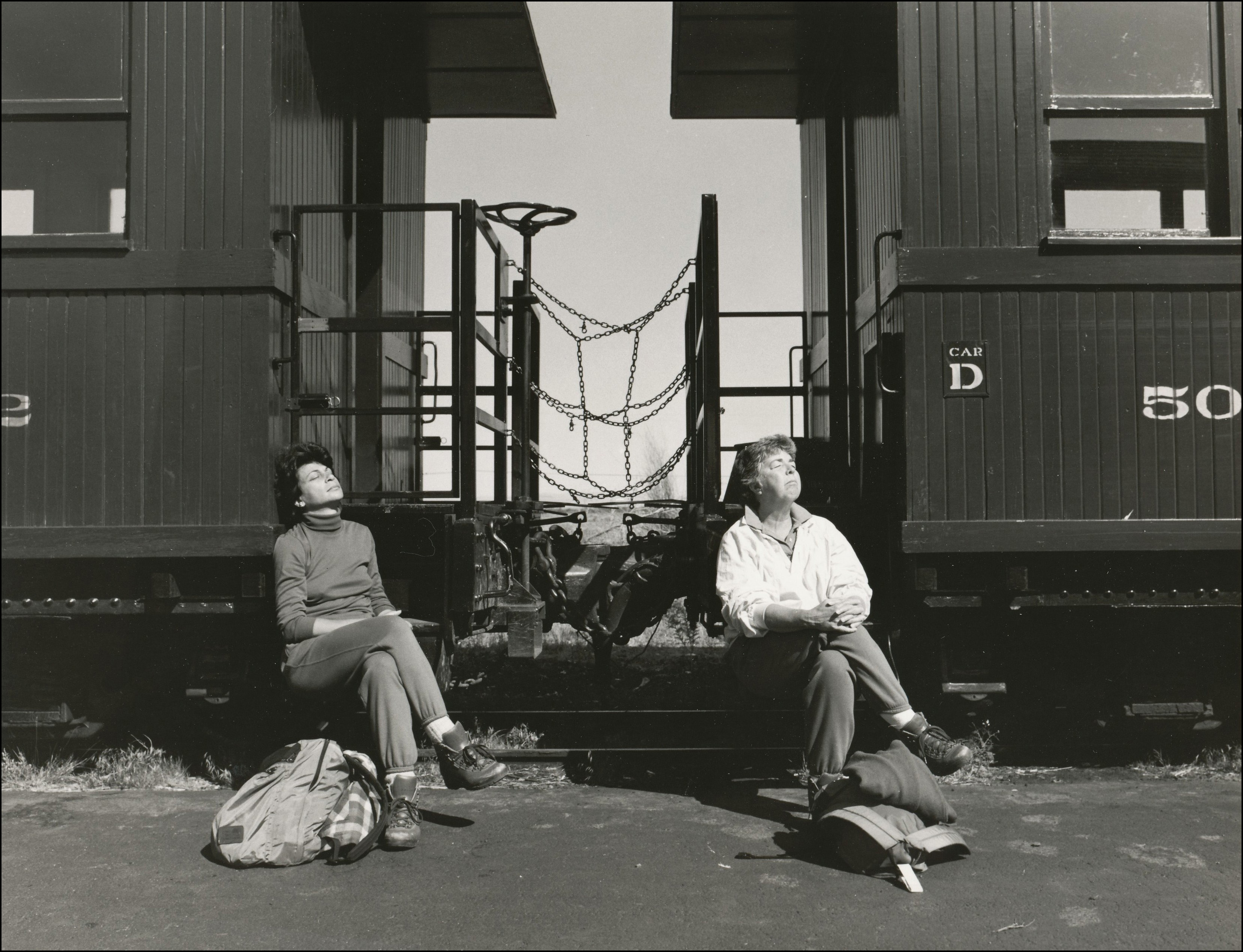 Two women waiting for a train. Sitting on the edge of the car stairs with eyes closed & faces pointed up towards the sun, legs crossed and their bundles of belongings at their feet.
