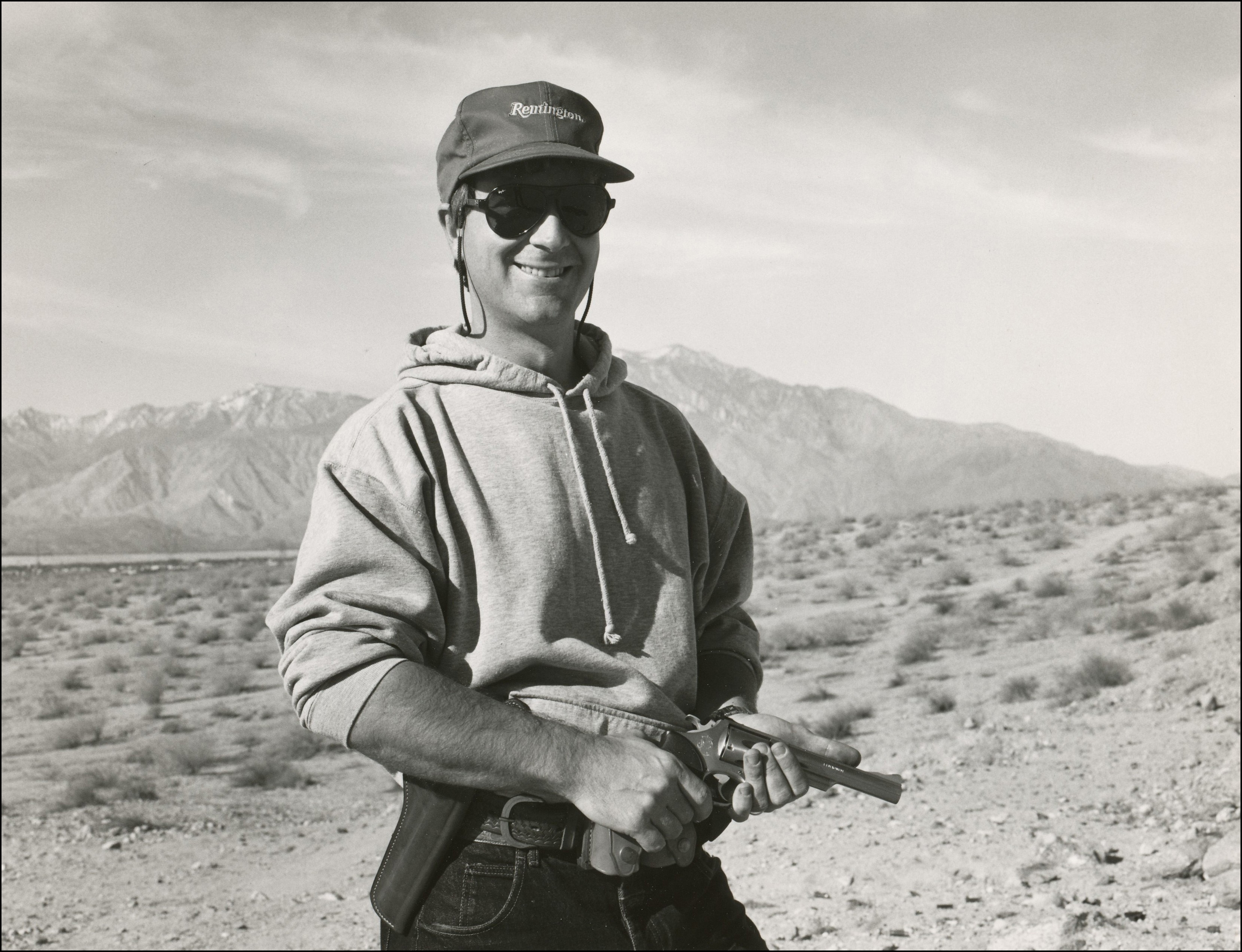 Man in baseball cap, sunglasses and hoodie standing with pistol in hand. In dessert area with mountains in the background