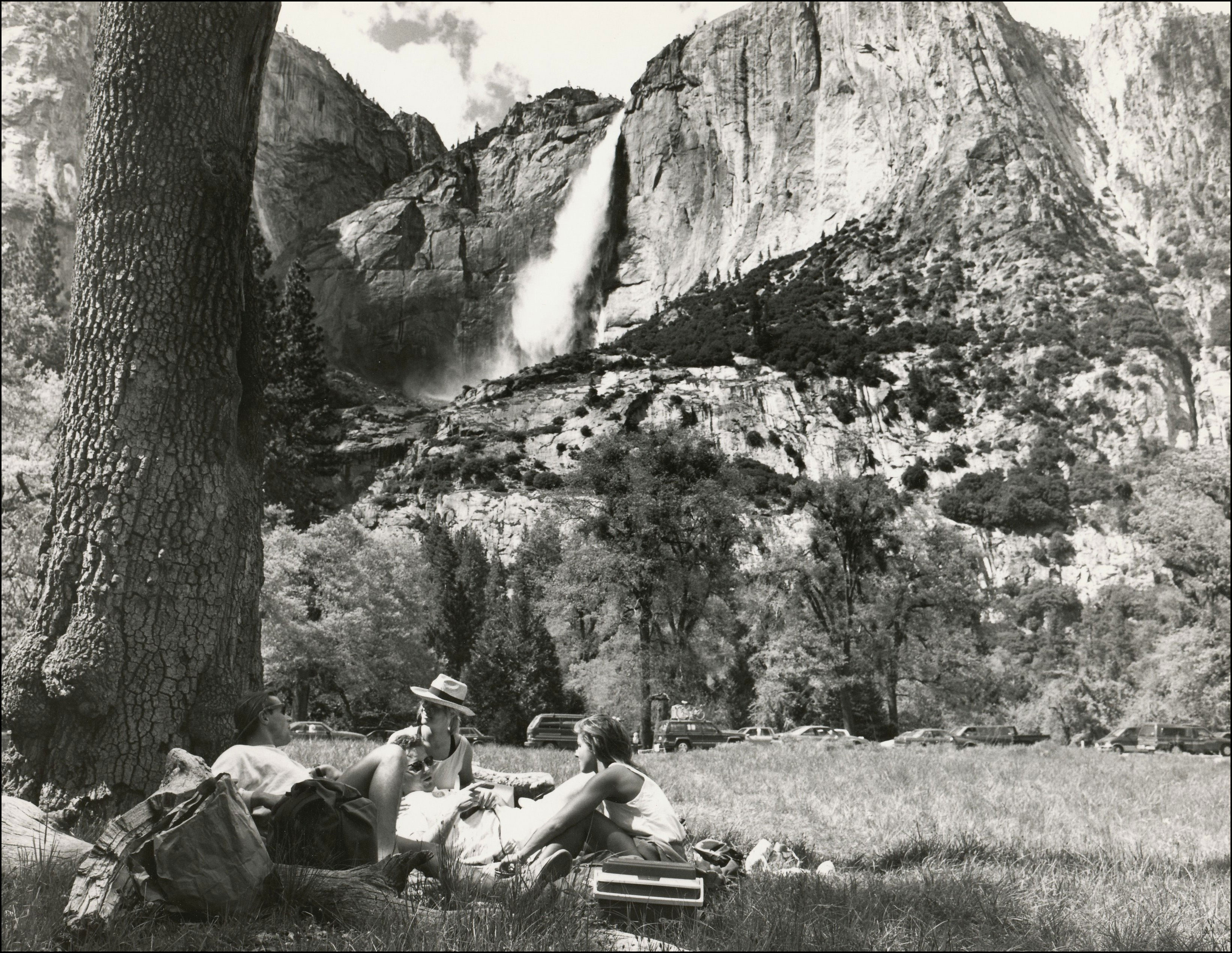 Two people sitting under a big tree having a picnic. View of parking lot in front of mountain with waterfall