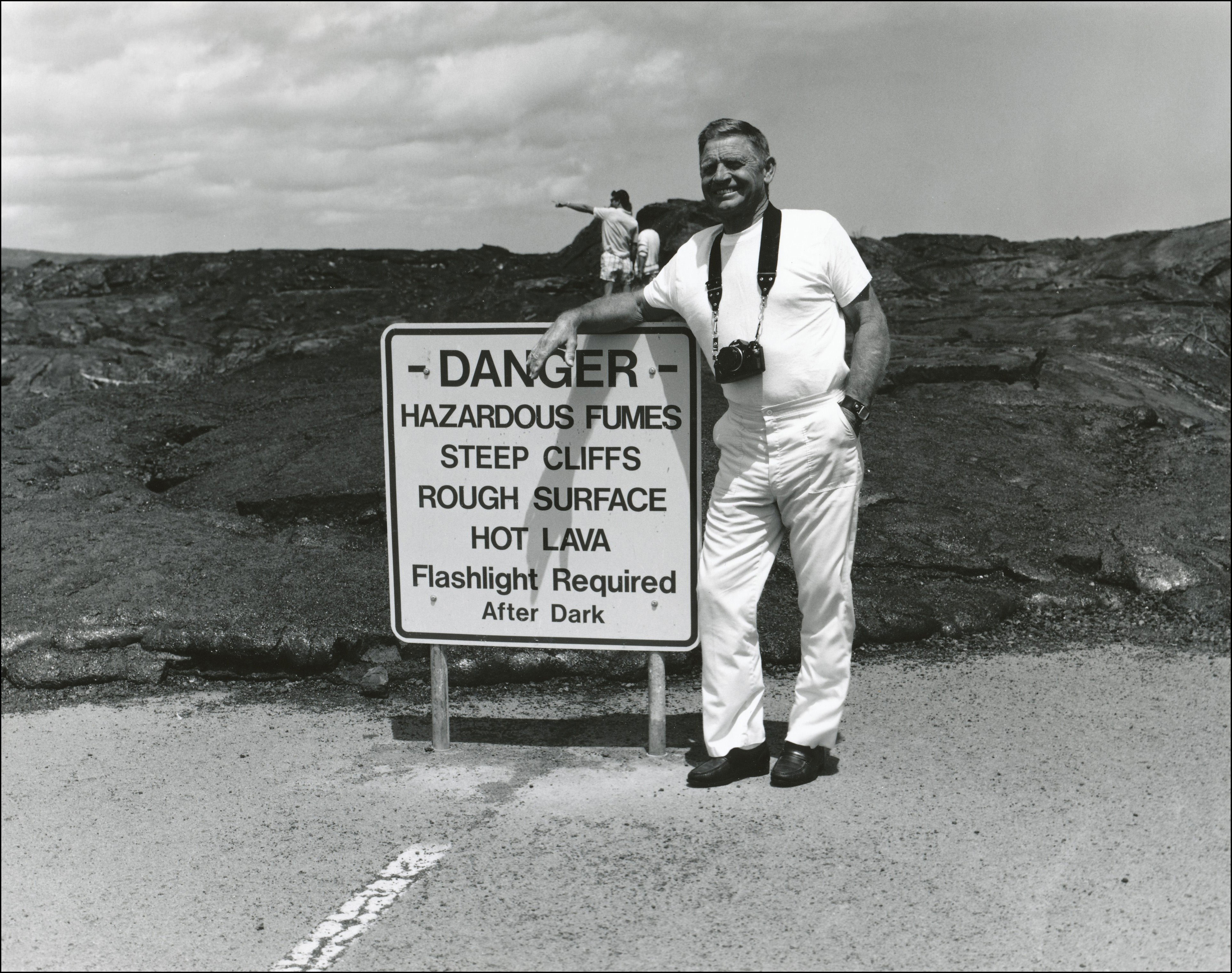 Tourist with camera around neck posing by a sign that says DANGER, hazardous fumes, steep cliffs, rough surface, hot lava, Flashlight required after dark. Old lava flows behind him.
