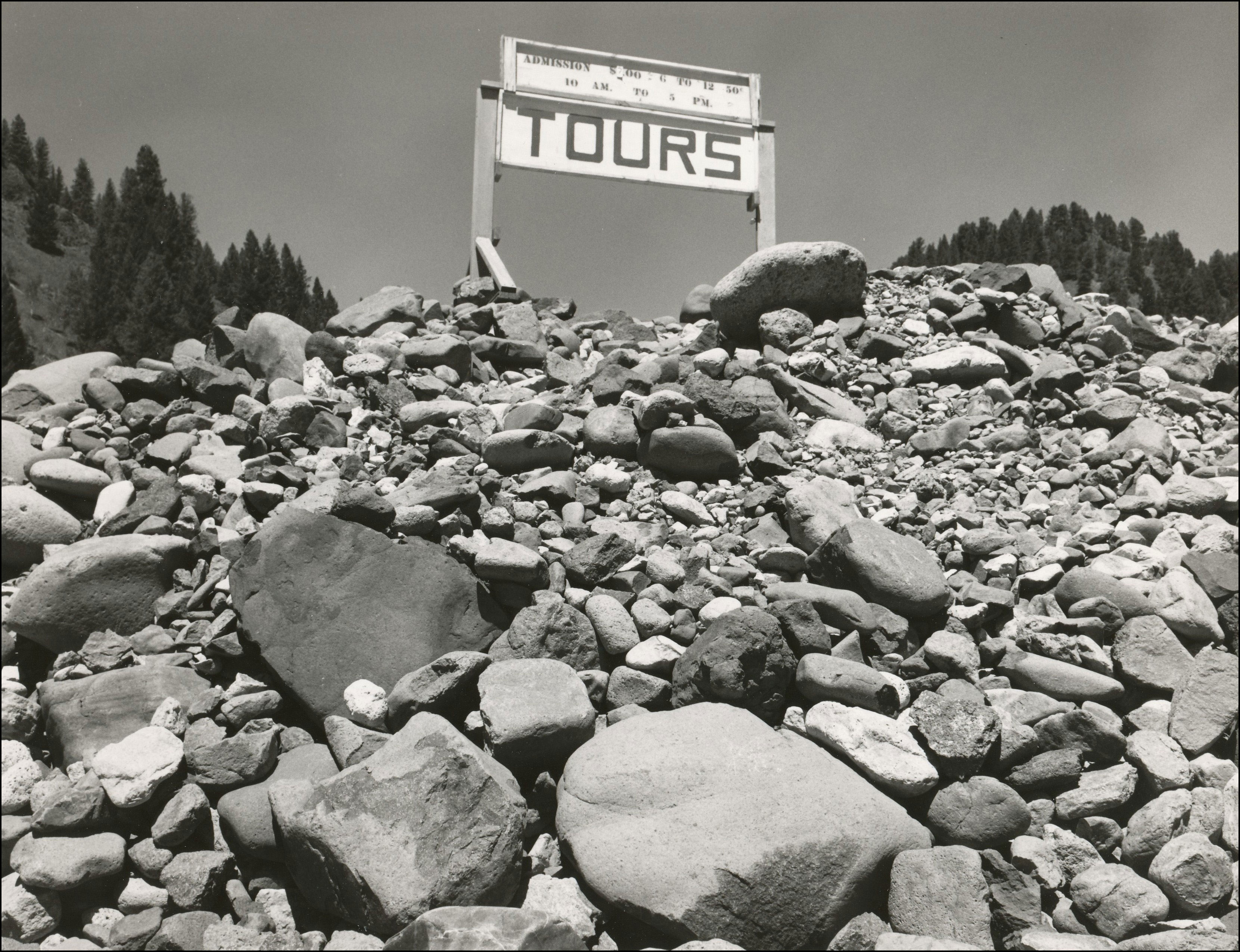Looking up at a sign that says TOURS with times above. The sign is on a large pile of rocks of all sizes.