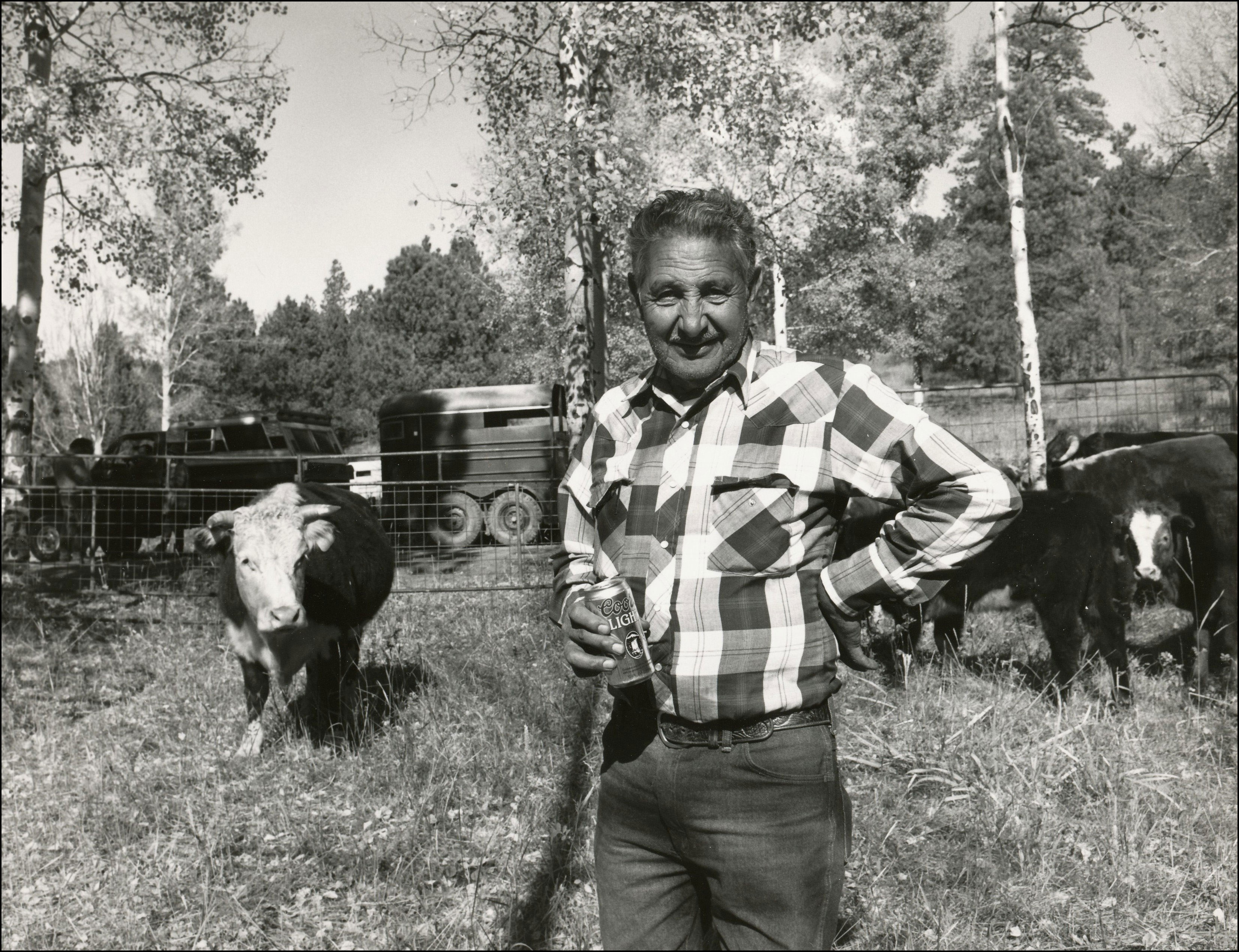 Rancher standing in pasture holding a can of coors light with hereford cattle behind him.