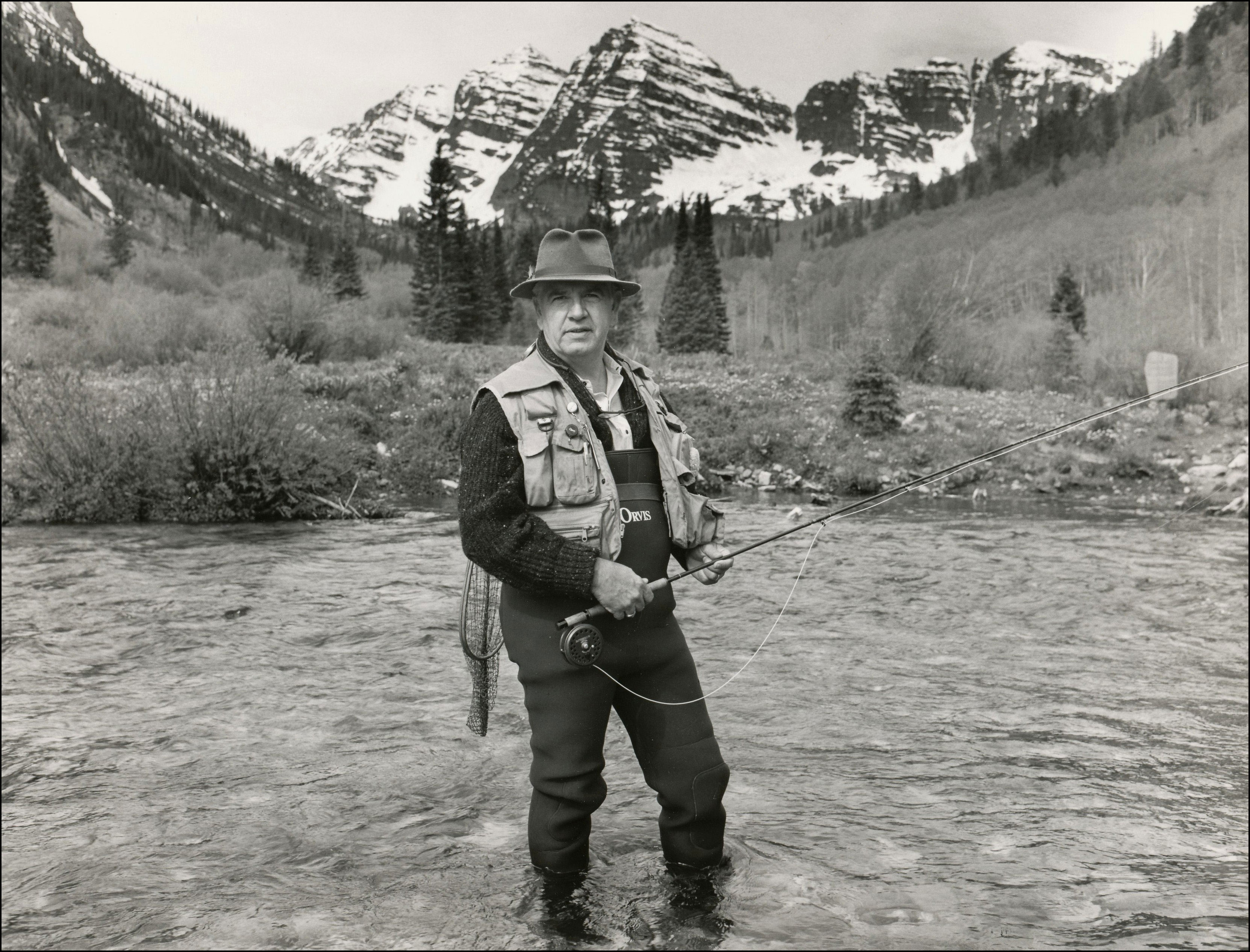 Fisherman with hat, vest and waders standing in river with fly rod. Rugged mountain peaks with snow in the background.