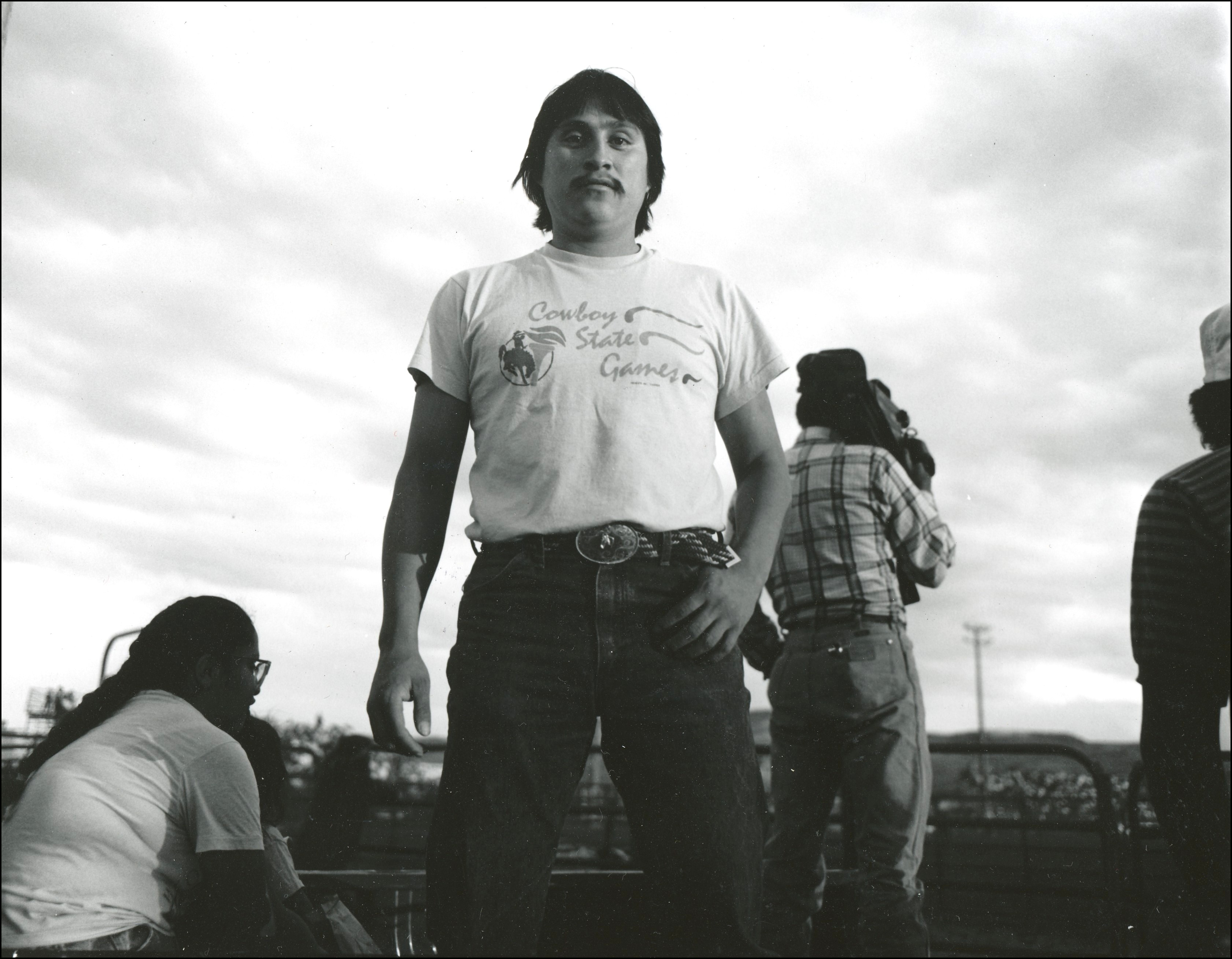 Native cowboy standing, wearing a t-shirt, western belt and buckle, and posing for camera, rodeo arena behind him. person sitting to his right and one standing with a video camera to his left.