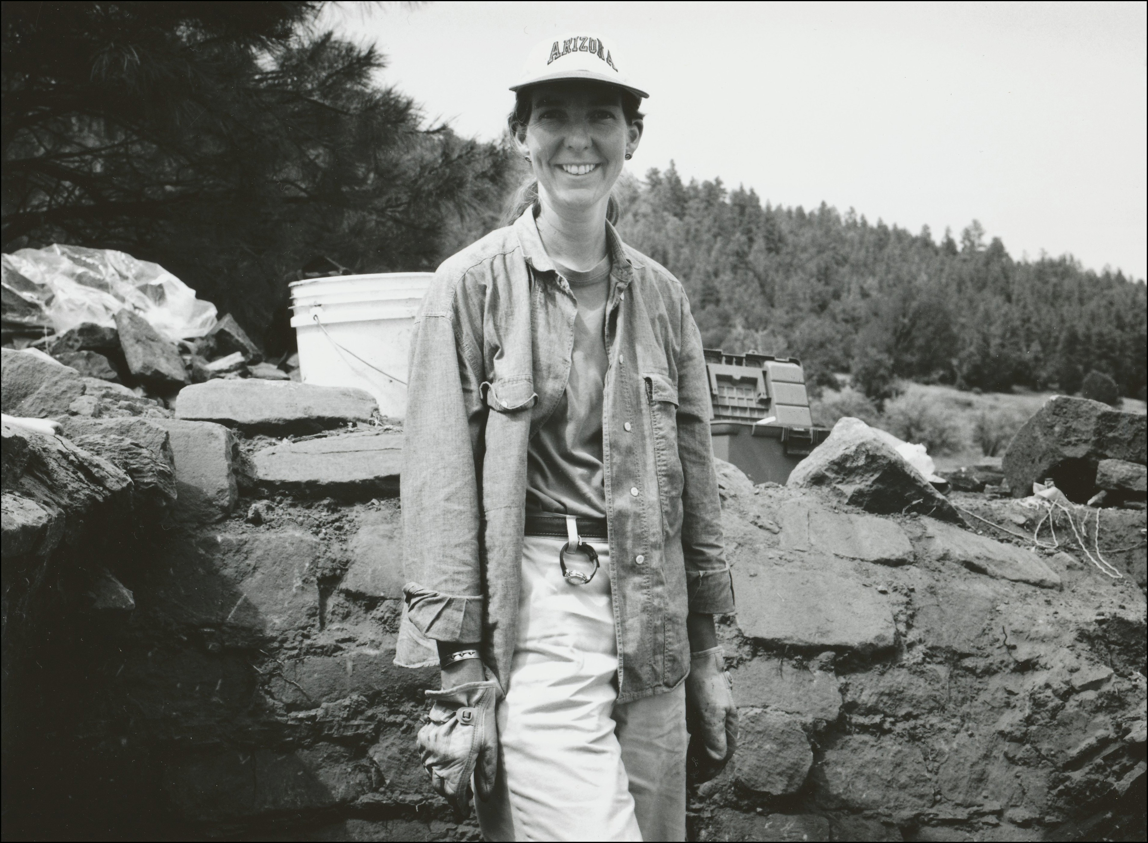 archaeologist with cap and gloves posing in front of old stone wall with bucket and toolbox in the background.