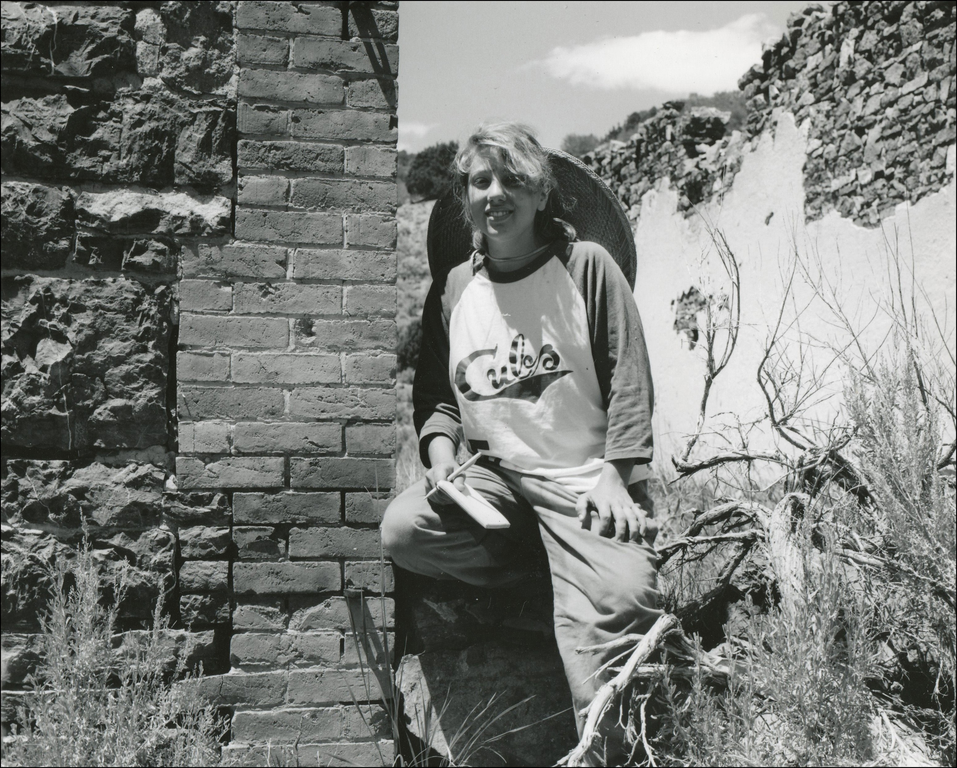 Young woman with Cubs shirt, notepad and pen leaning with one leg up against historical brick building