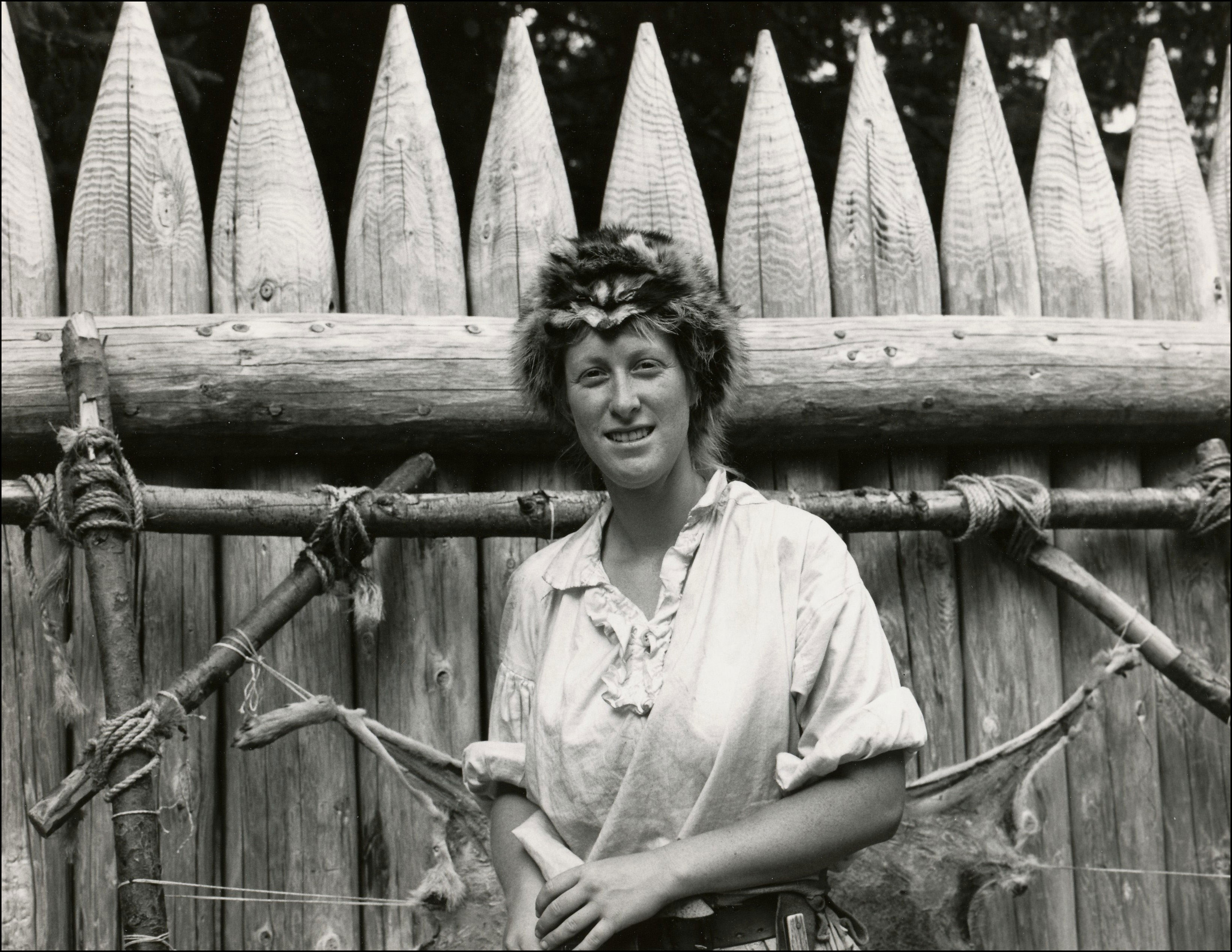 Woman with coon cap and mountain-main attire standing in front of an old fort type fence. Hide if an animal is stretched out to dry behind her.