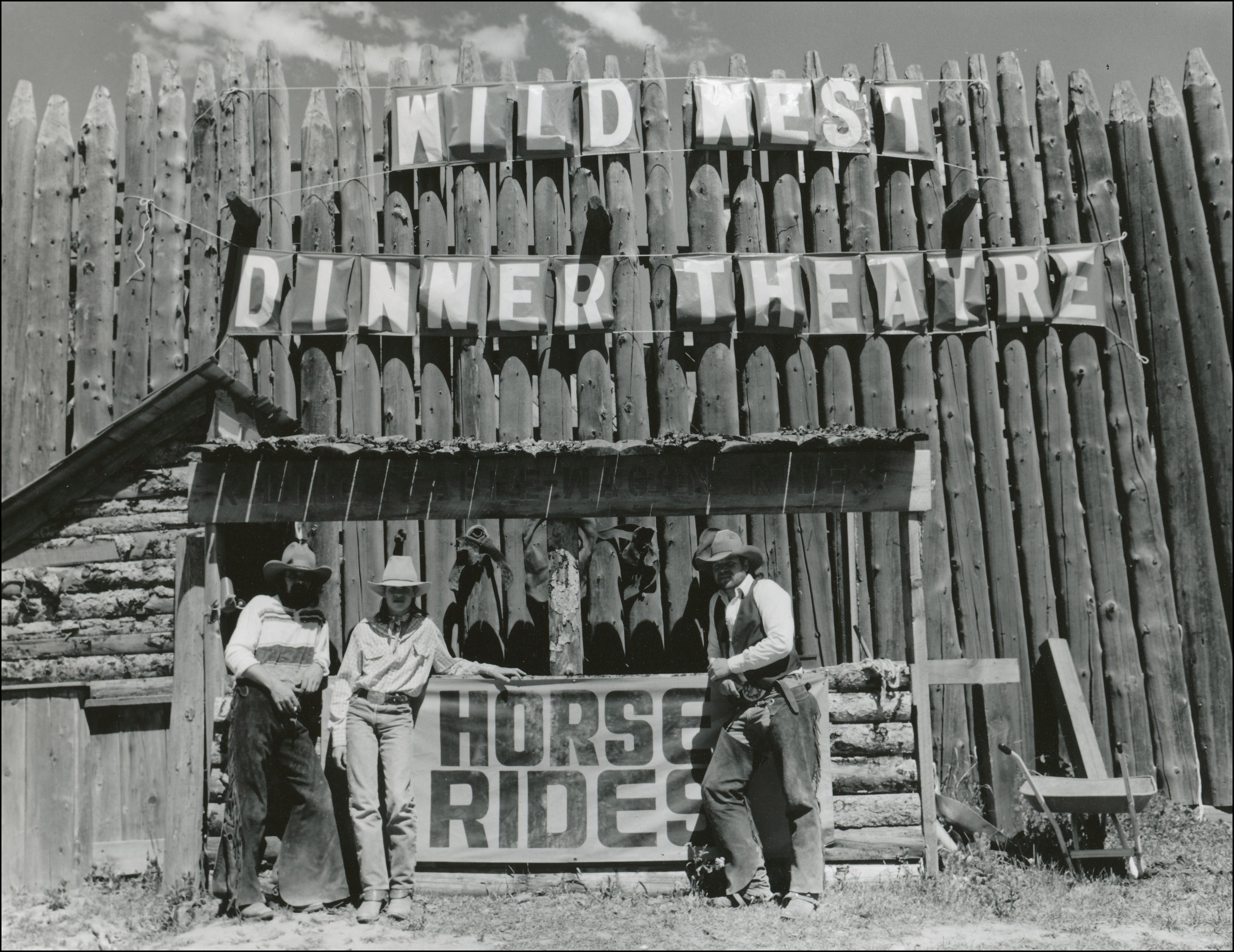 Three people in western wear standing in front of for fence with sign above that says Wild West Dinner Theatre and a booth that has a sign that says horse rides