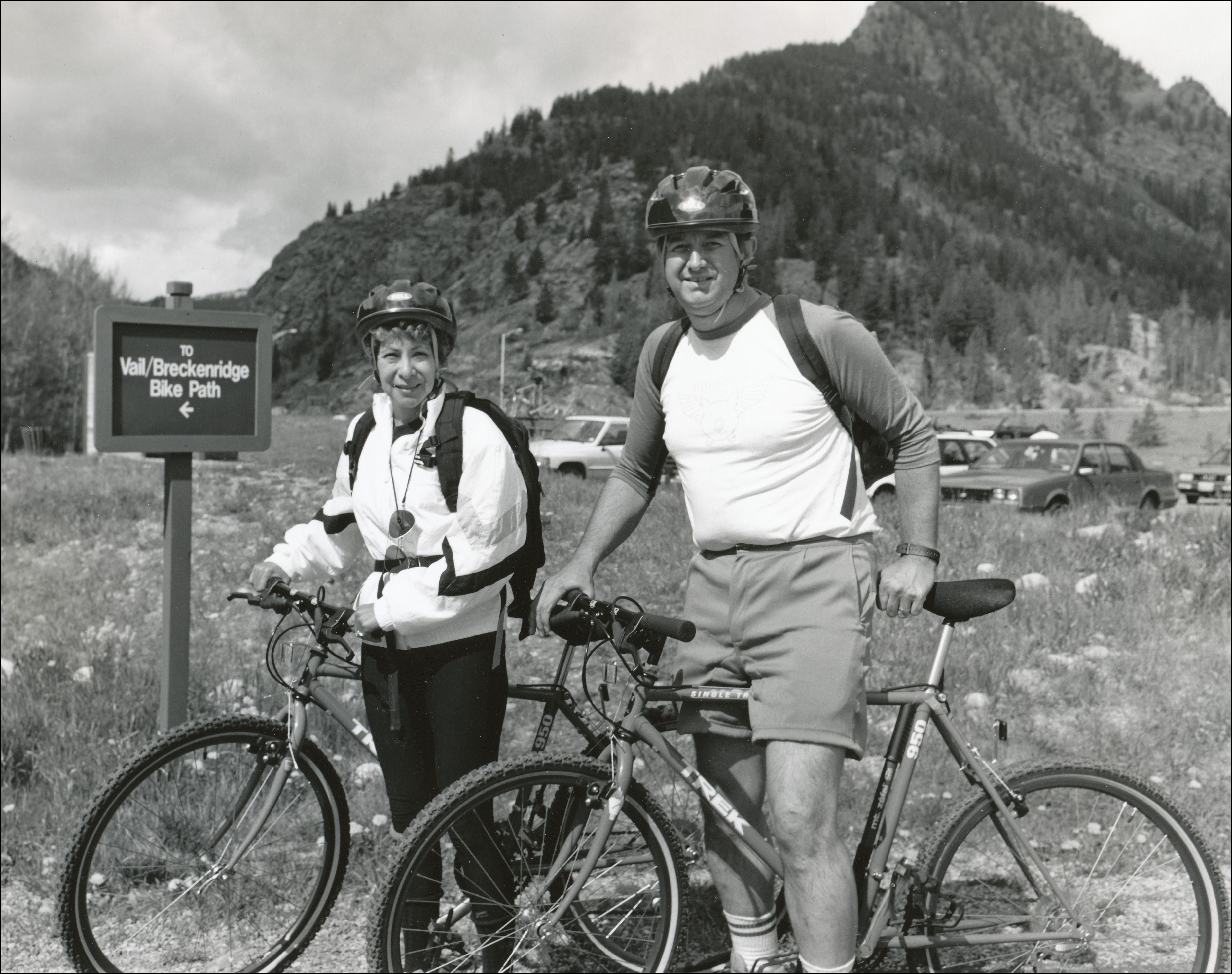 Two mountain bikers posing for the camera. Parking lot with cars behind them and sign to their left which says to Vail-Breckenridge Bike Path