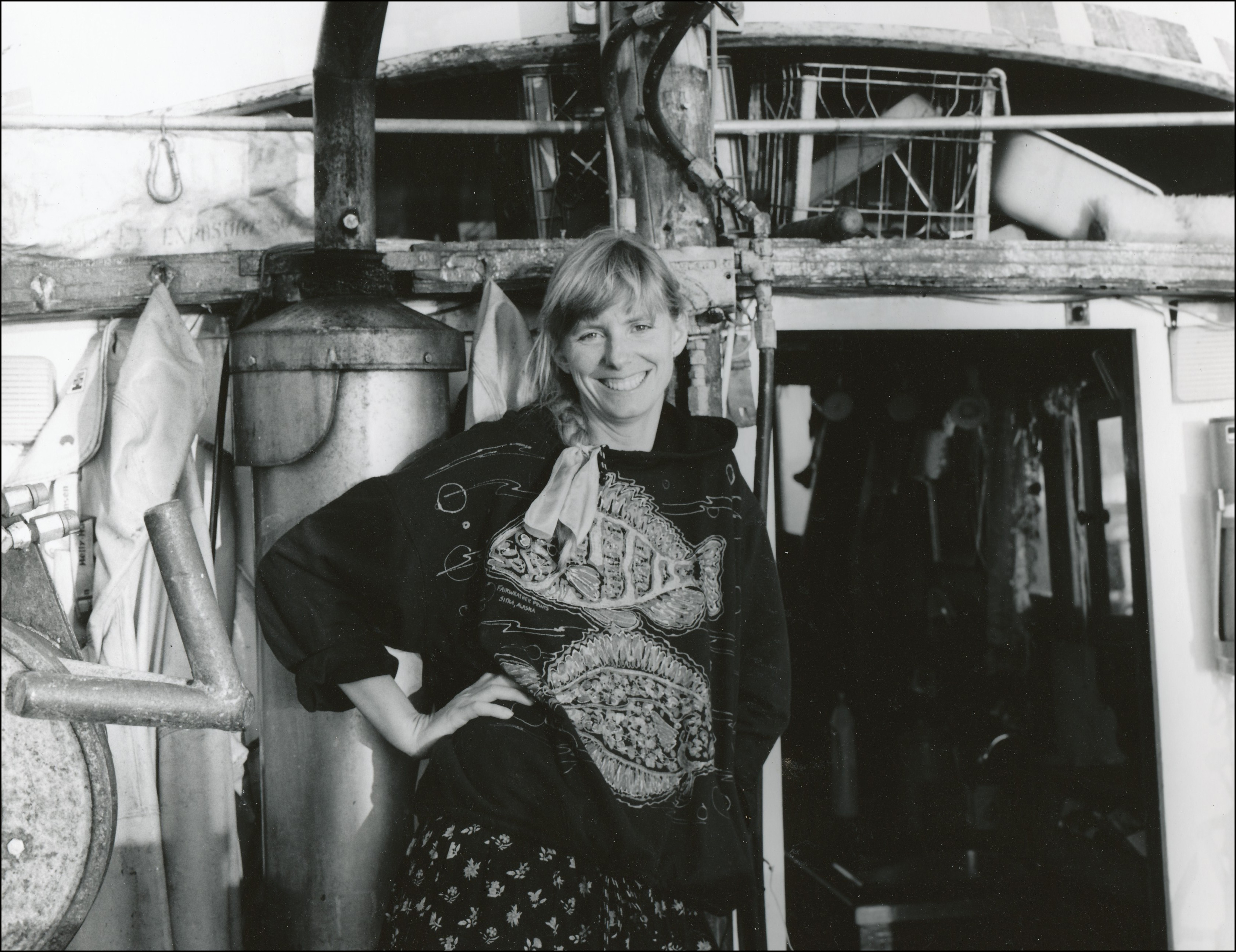 Woman standing in front of entrance to the cab of a boat smiling for the camera on hand on hip. She is wearing a sweater that has a picture of the anatomy of a fish. 