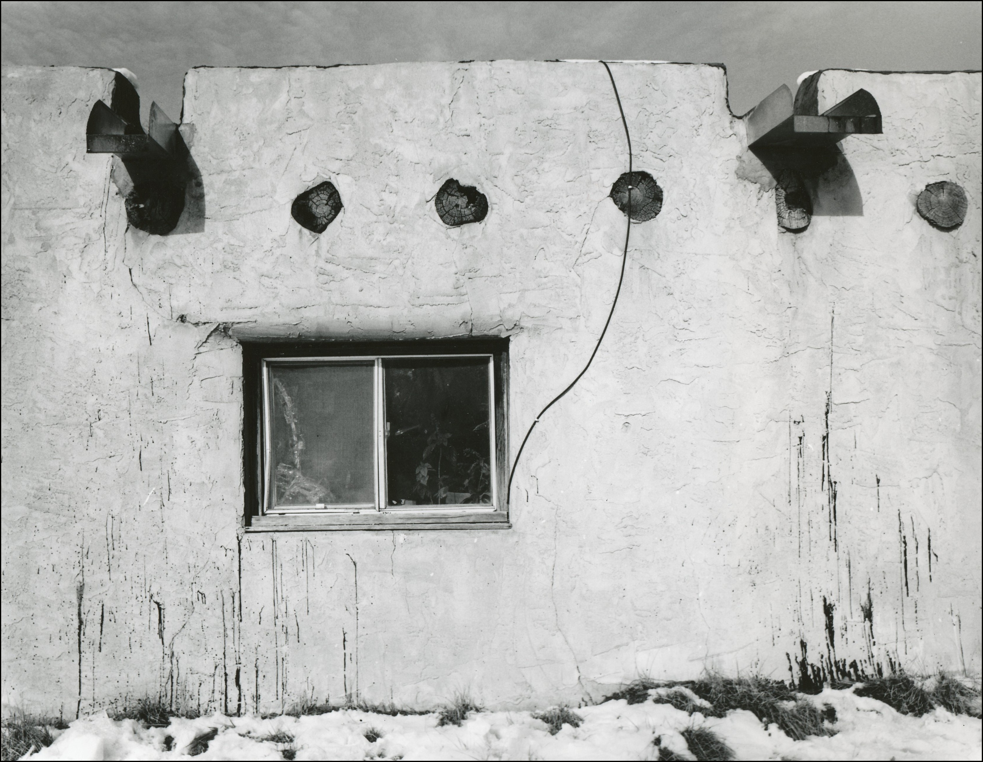 Side of adobe home with window, beems and rain gutters. Snow on the ground in front.