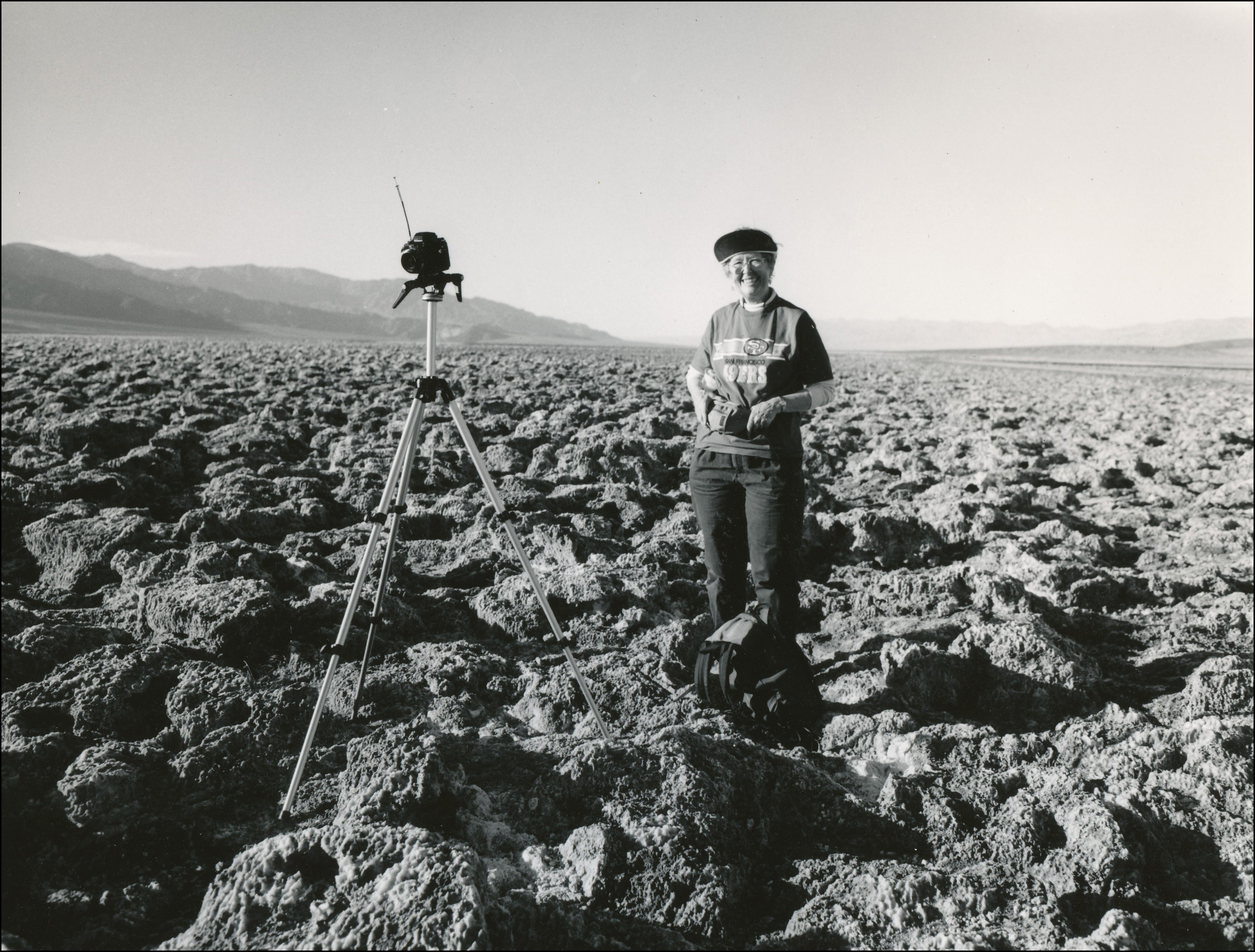 Woman standing next to camera on tri-pod in the middle of a flat in the mountains that has large rocks all over