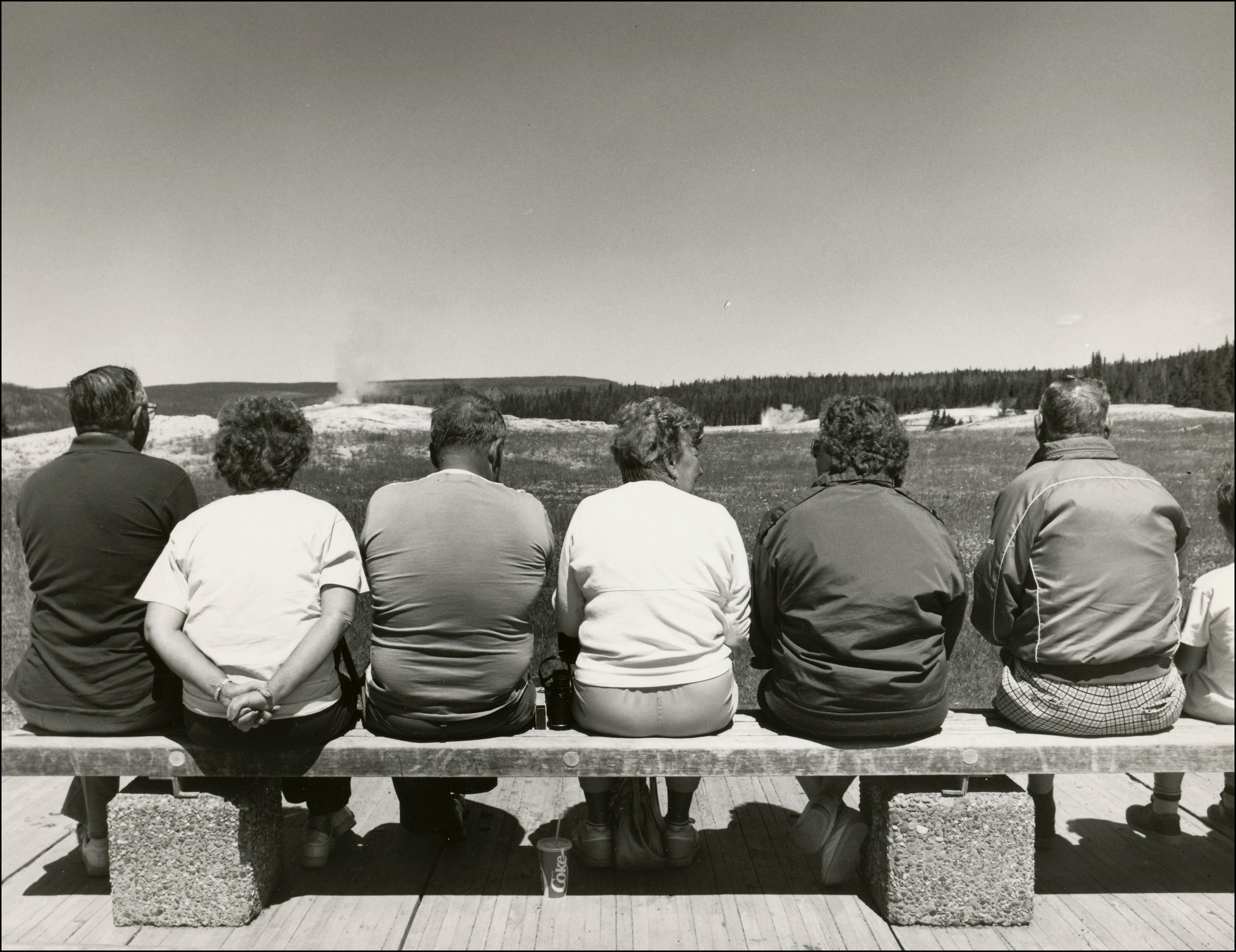 View of the backs of 6 people sitting on a bench watching steam from a geyser