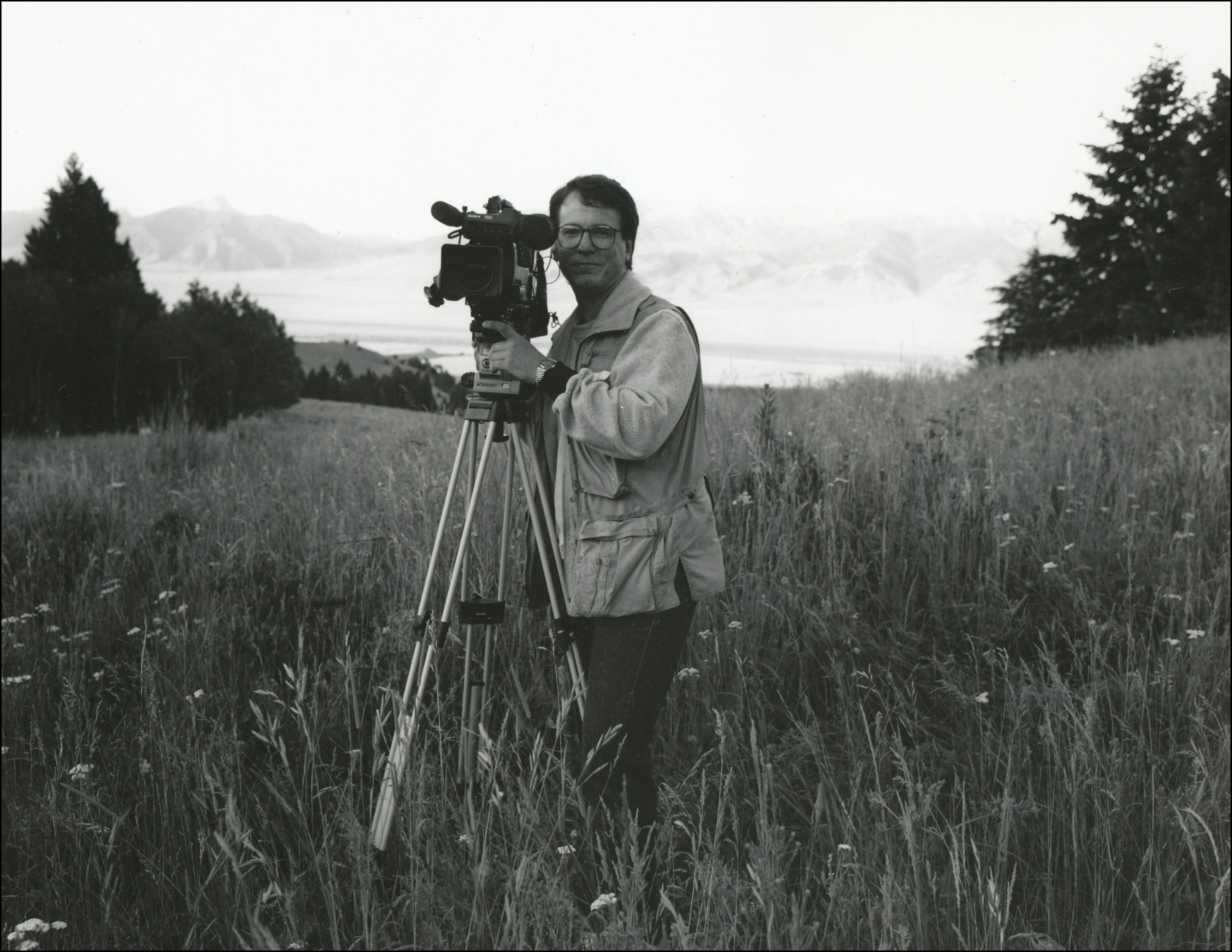 Man standing with his camera on a tripod in an open grassy area with mountains in background