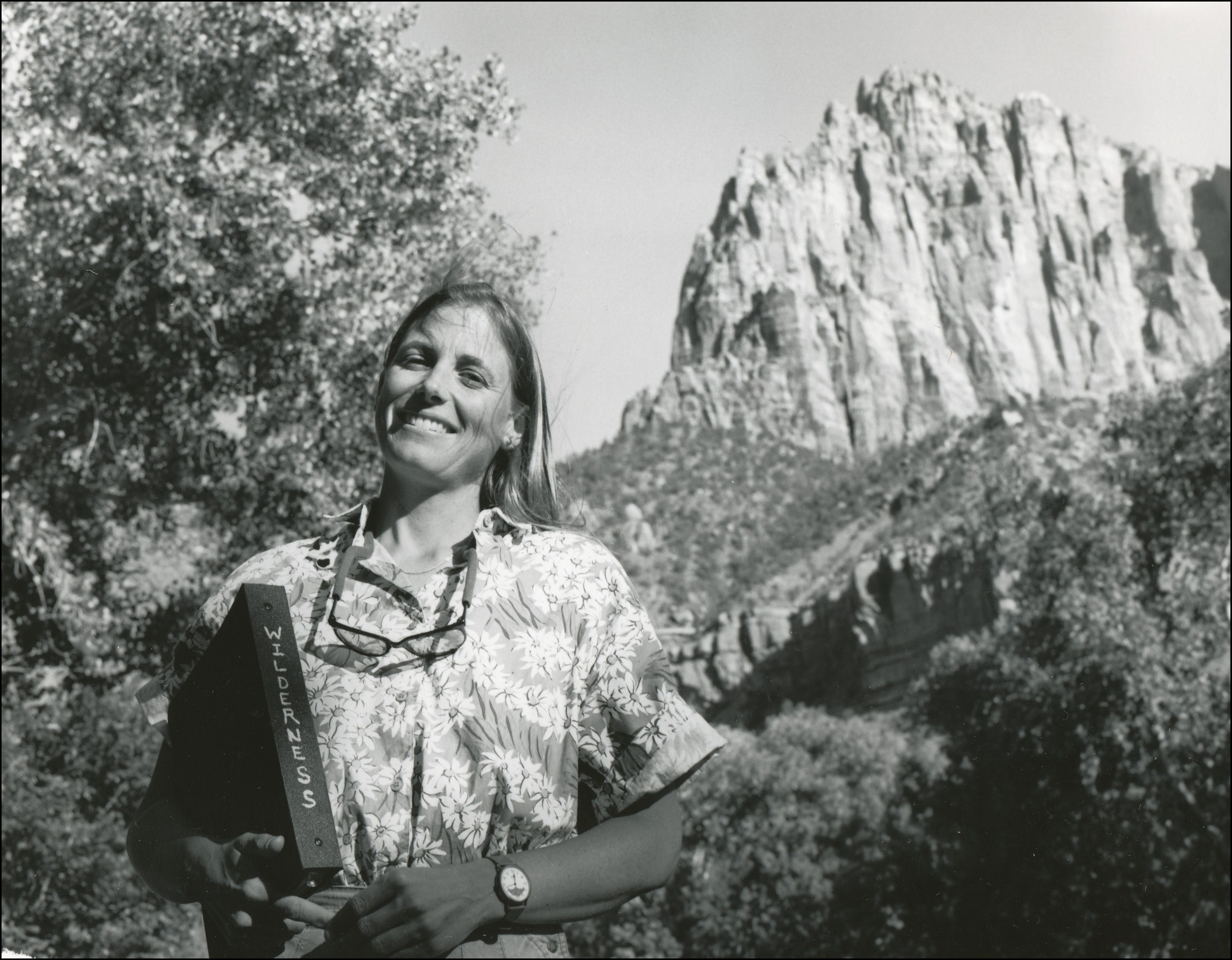 Woman smiling with glasses hanging around neck holding a book labeled wilderness with rocky peak in the background