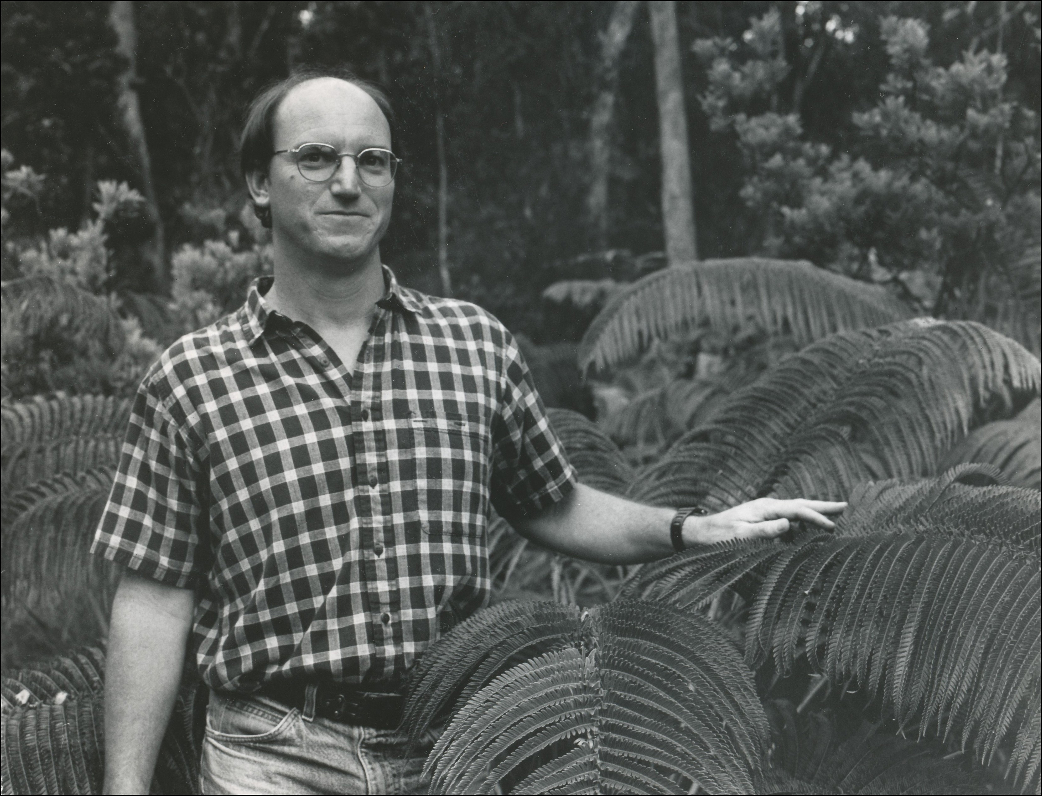 Man with glasses in short sleeved button up plaid shirt standing close to large fern with his left hand touching large fern leaf