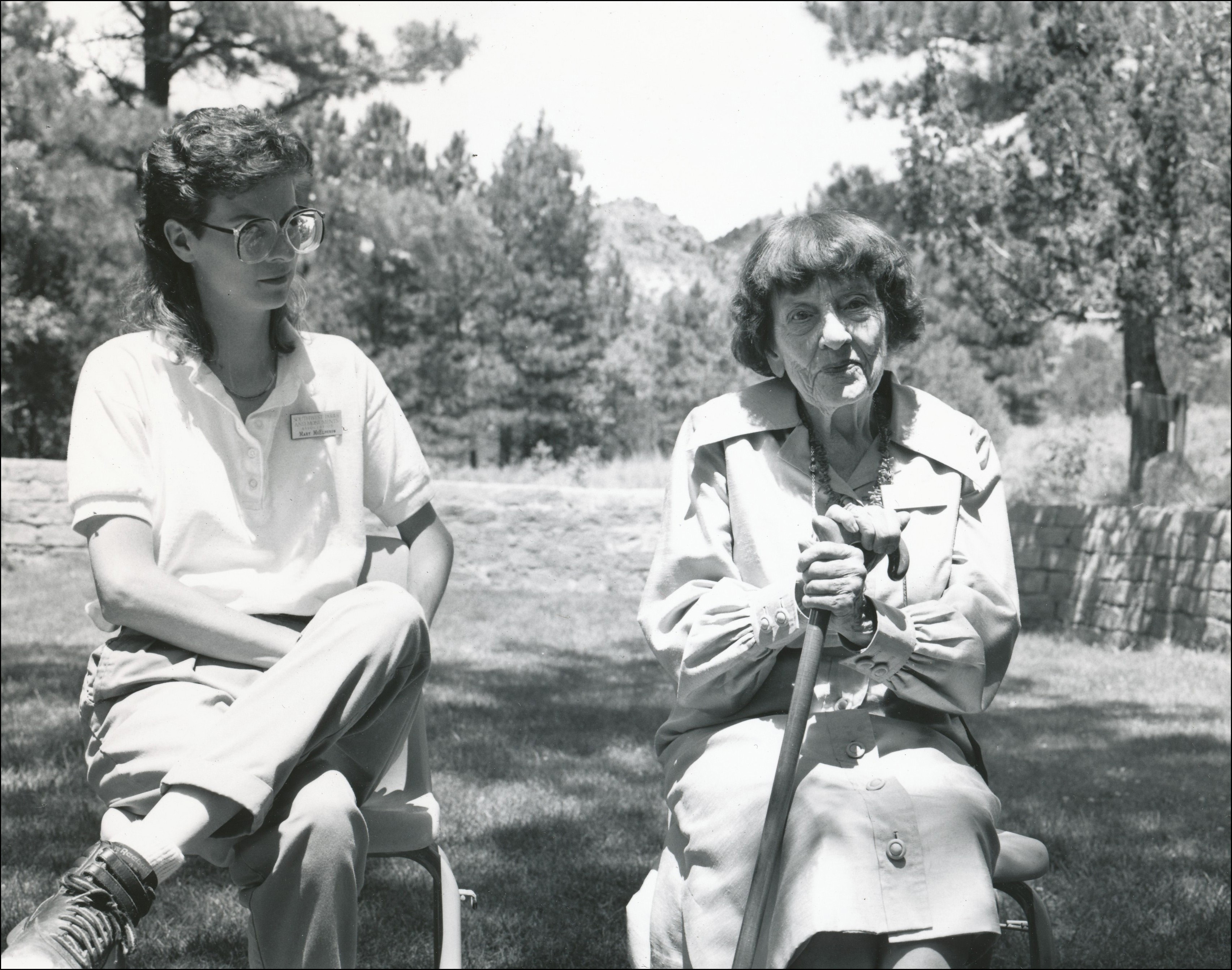 Two women sitting outside on chairs with ponderosa pine in the background. Woman on left is younger and woman on right is older holding her cane