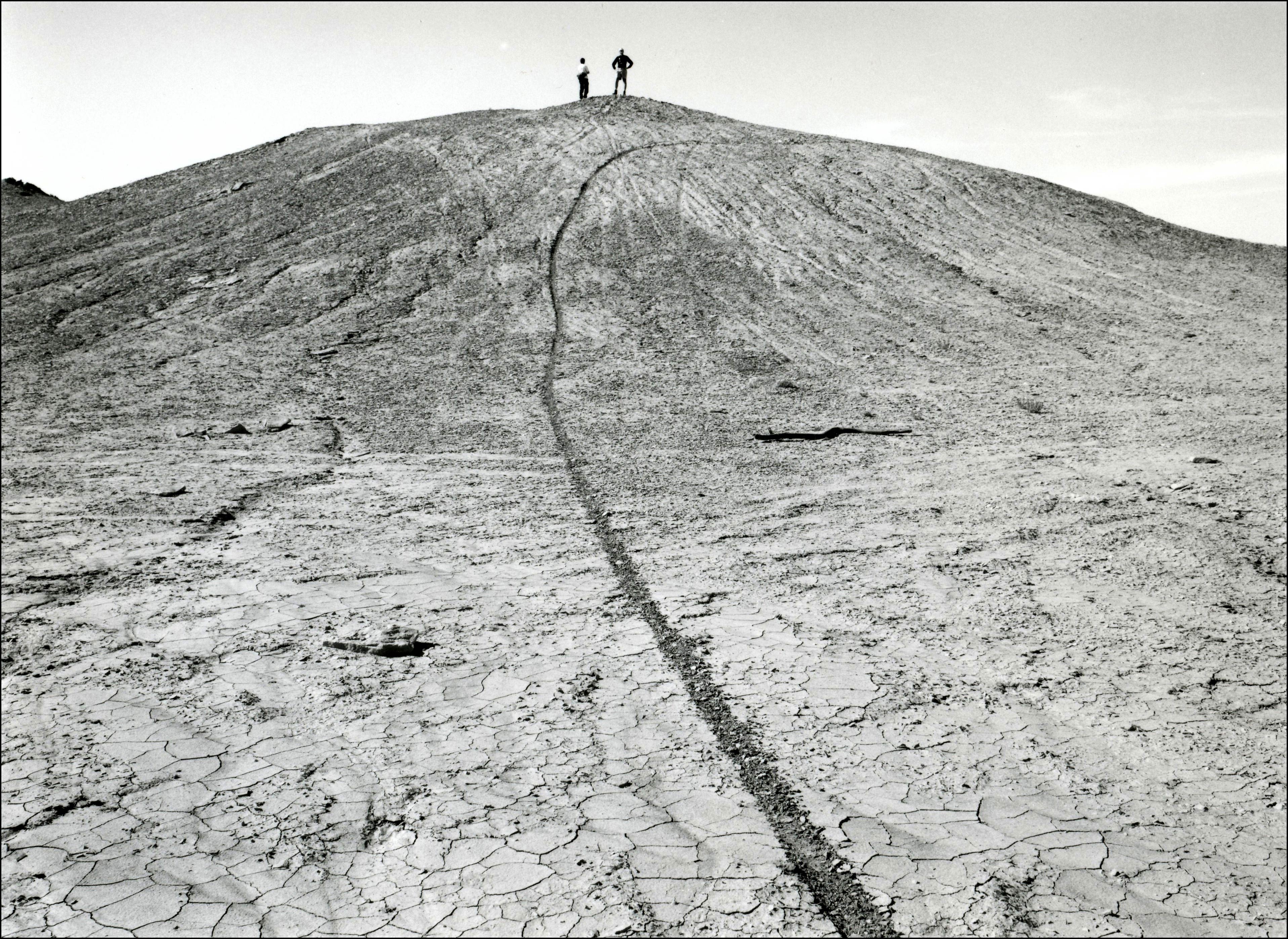 Two people standing on top of a hill in an area with cracked dirt and no vegetation. There is a single dirt bike track going up the hill.