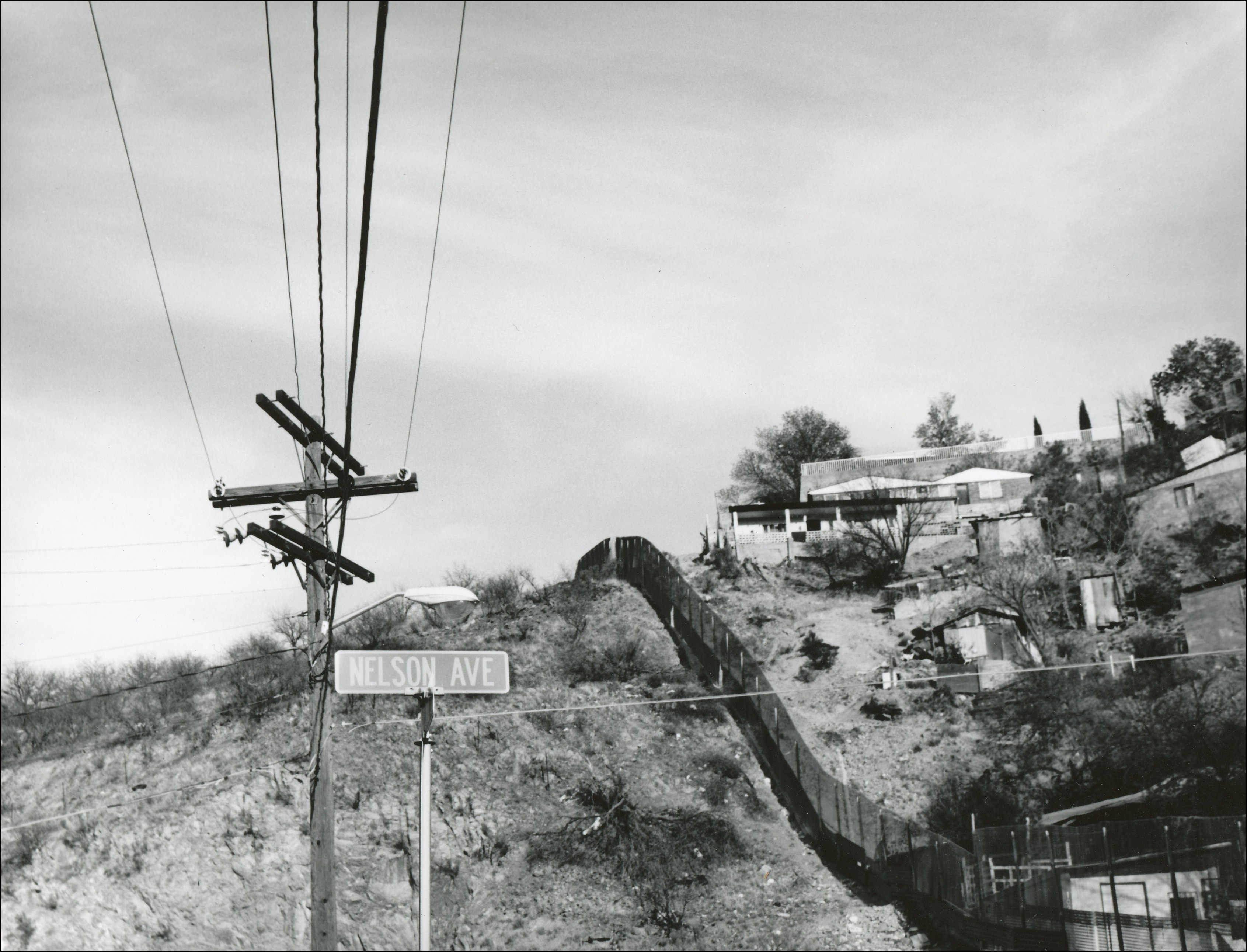 View of a border fence along a hills. Houses on right side and vegetation on left side. Also view of power lines and street sign that says Nelson Ave