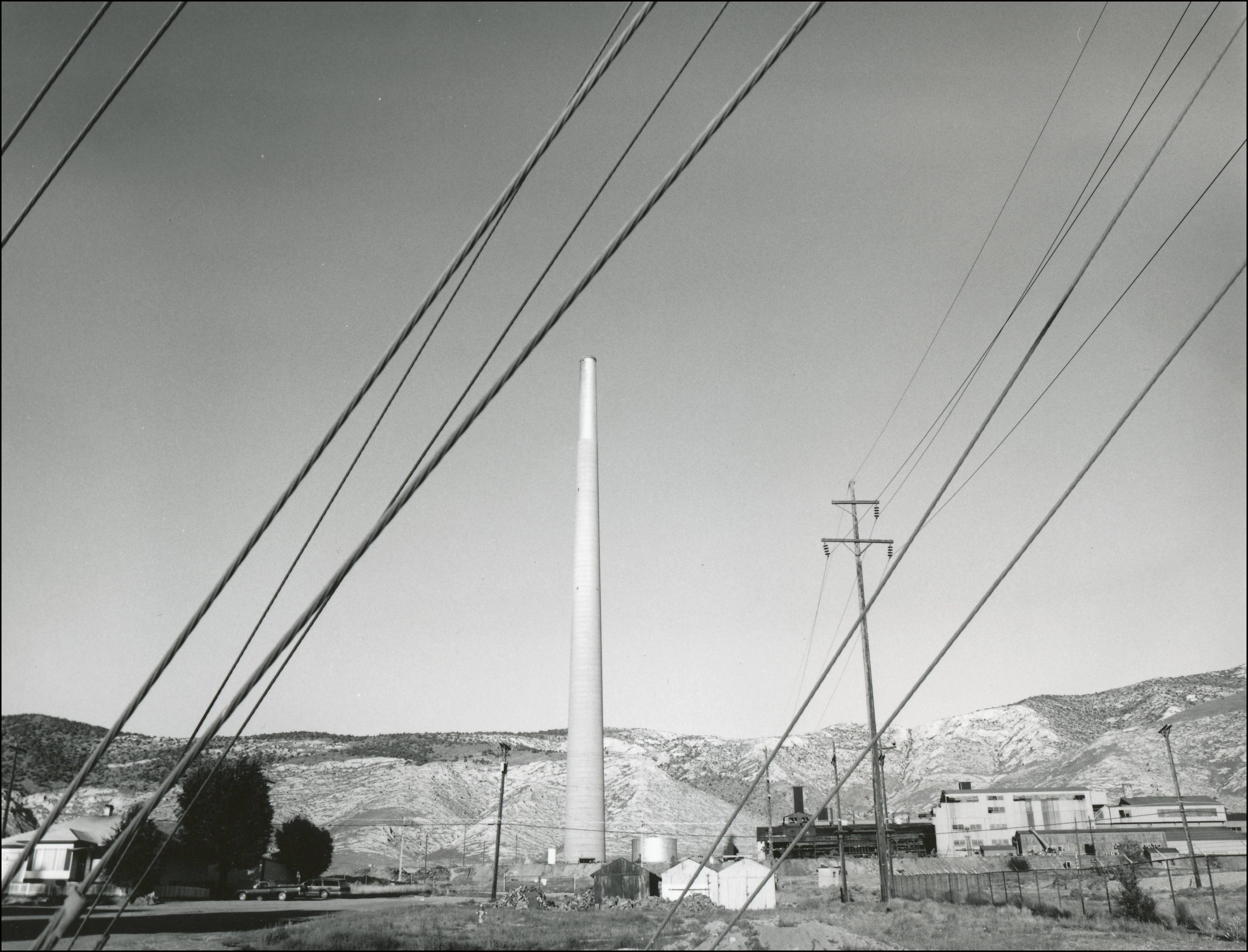 View through power lines of tall smokestack. A few houses and buildings are in surrounding area.