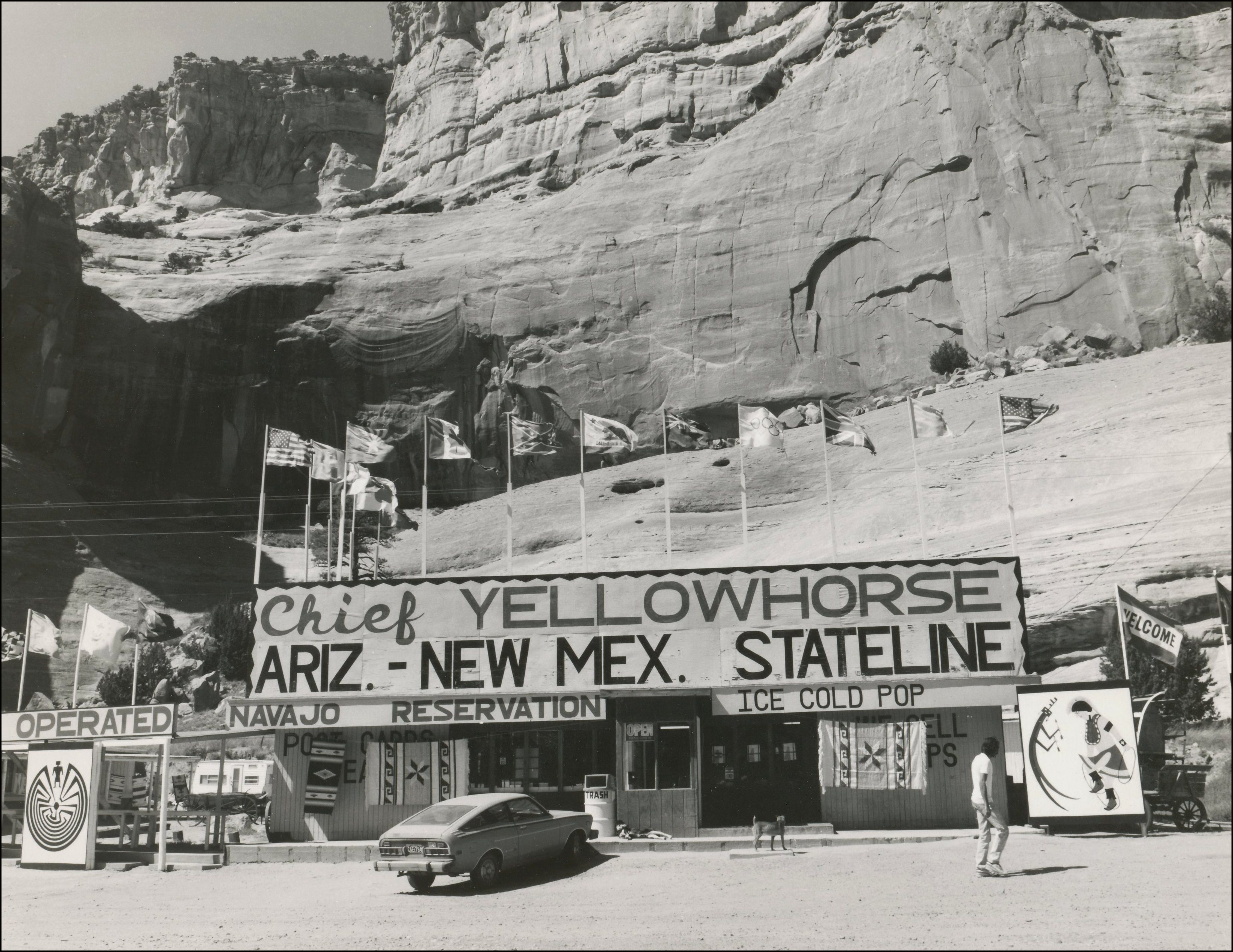 Road side stop store in foreground with flags and says Chief Yellowhorse, Ariz- New Mex. Stateline, Navajo reservation, ice cold pop. Large cliffs behind it.