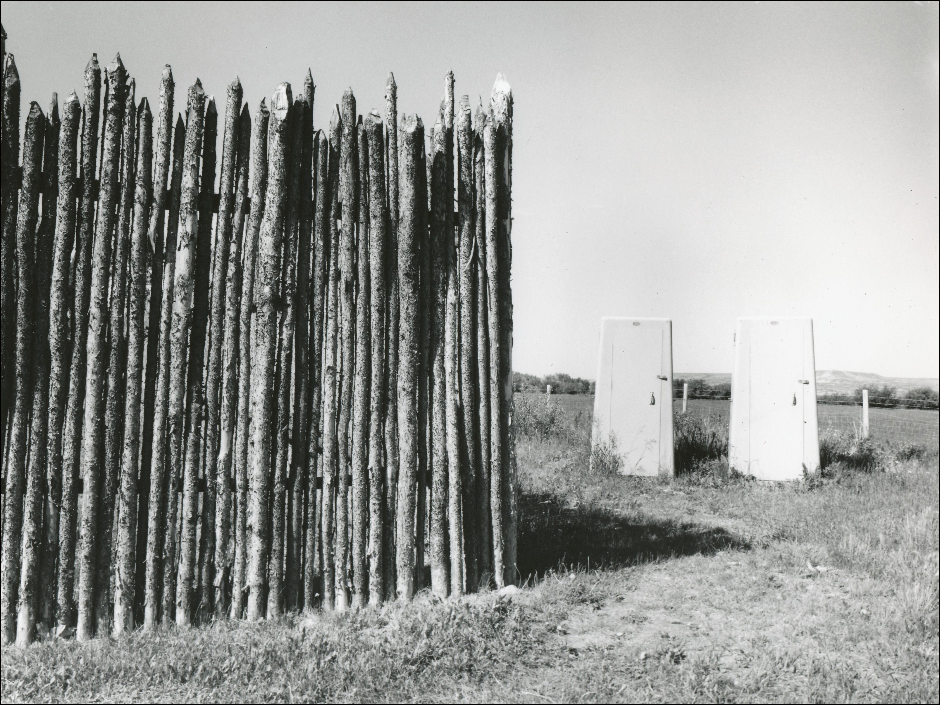 2 portapotties behind an old fort fence made of lodgepoles with pointed tops
