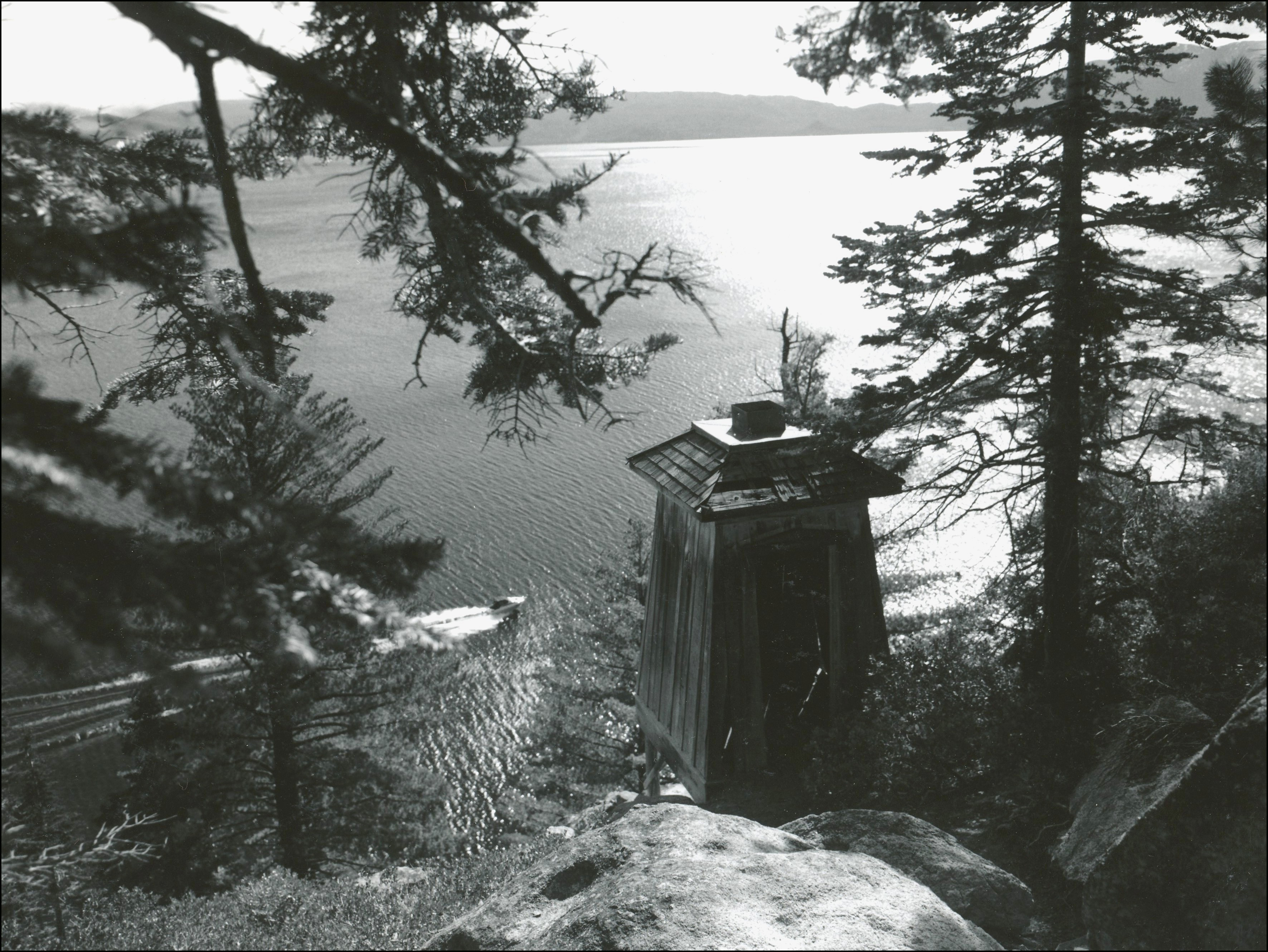 View of lake through the trees on the shore. Closer to the lake up on rock ledge is an old lighthouse and down below on the lake is a motor boat