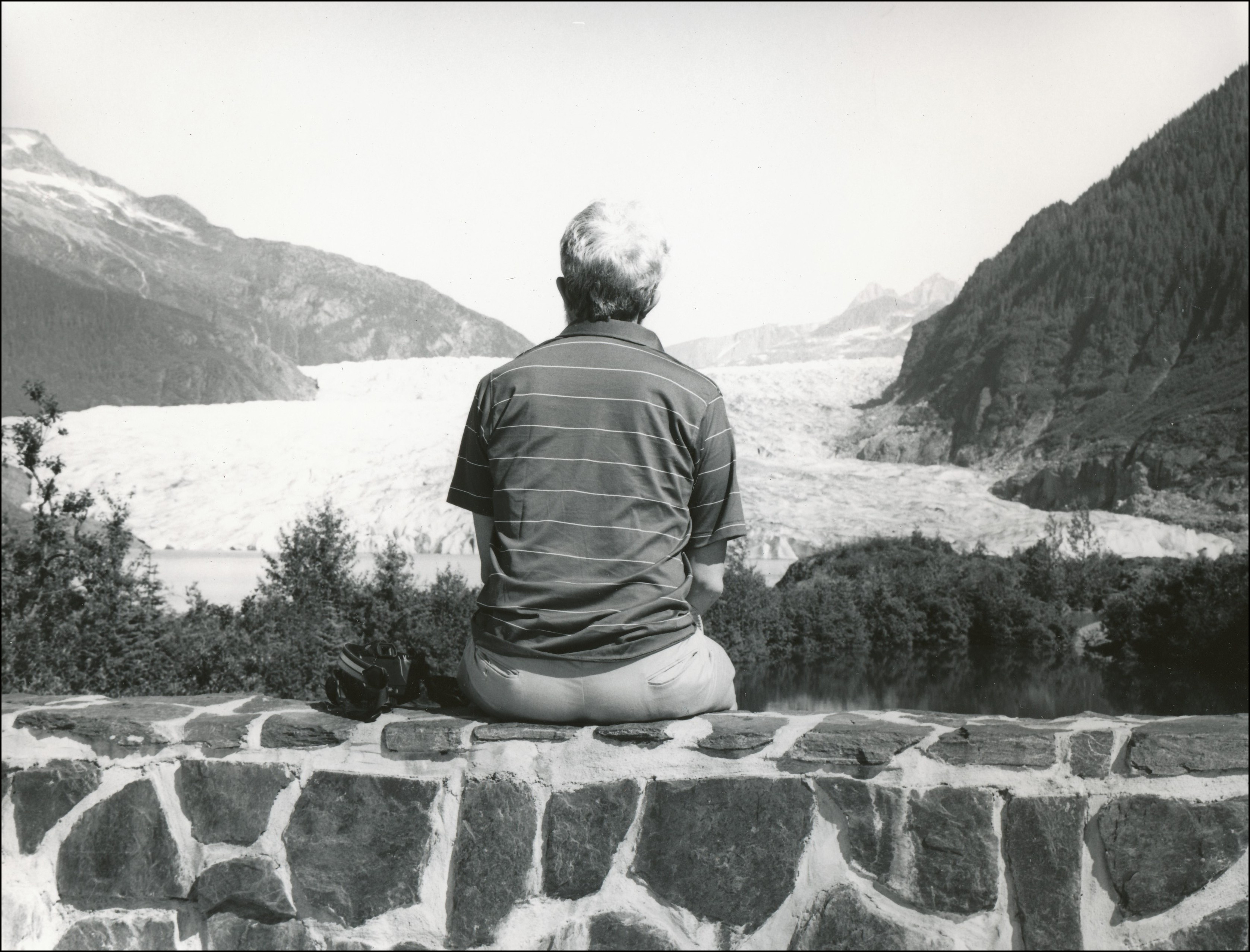 Man sitting on stone wall looking out at large glacier inbetween mountains